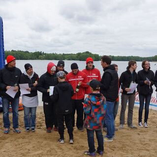 Gruppenansprache von Teilnehmern mit Urkunden und Medaillen am Strand eines Gew&auml;ssers bei bew&ouml;lktem Himmel.