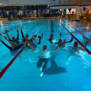 Gruppe von Schwimmern mit erhobenen H&auml;nden in einem beleuchteten Hallenbad mit blauer Wasseroberfl&auml;che.