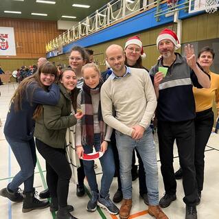 Gruppe von acht Personen, einige mit Weihnachtsm&uuml;tzen, beim Feiern in einer Sporthalle mit festlicher Dekoration.