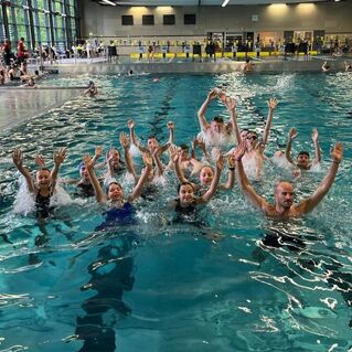 Gruppe von Schwimmern im Wasser, die l&auml;chelnd die H&auml;nde heben, umgeben von einer Poollandschaft.