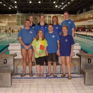 Gruppenfoto von acht Schwimmern in blauen Shirts vor einem Schwimmbecken mit Startbl&ouml;cken und Wettkampfatmosph&auml;re.