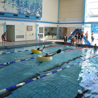 Schwimmhalle mit Teilnehmern in aufblasbaren Ringen, w&auml;hrend Betreuer am Beckenrand zuschauen.