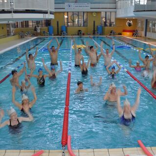 Gruppenschwimmen mit zahlreichen Jugendlichen, die im Wasser stehen und ihre Arme heben, in einem Schwimmbad.