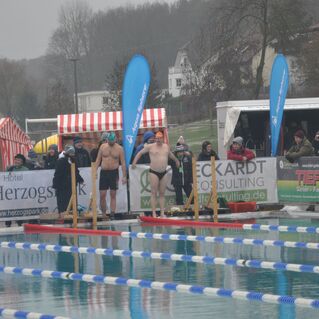Zwei Schwimmer stehen an der Startlinie einer Freiluft-Schwimmveranstaltung, Zuschauer und Banner im Hintergrund.