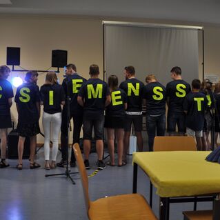 Gruppe von Menschen mit T-Shirts, die gemeinsam das Wort &bdquo;SIEMENSSTADT&ldquo; bilden, in einem Veranstaltungsraum.