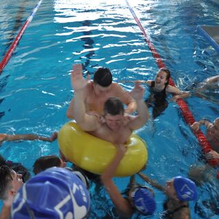 Gruppe von Schwimmern im Wasser, einer sitzt in einem aufblasbaren Ring, w&auml;hrend andere ihn anfeuern und unterst&uuml;tzen.