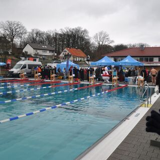 Wettkampf am Freibad, schwimmende Teilnehmer vor Zuschauern, umgeben von Zelten und einem k&uuml;hlen, bew&ouml;lkten Himmel.