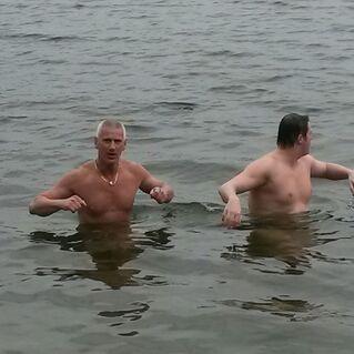 Zwei M&auml;nner ohne T-Shirts stehen im flachen Wasser eines Gew&auml;ssers und schauen in verschiedene Richtungen.