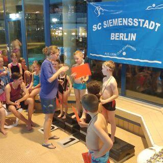 Gruppe junger Schwimmer steht an einem Startblock, w&auml;hrend ein Trainer Anweisungen gibt; im Hintergrund ein Vereinsbanner.