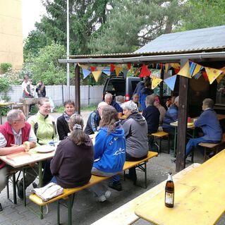 Gruppe von Menschen sitzt an Tischen unter einem &Uuml;berstand, farbige Wimpel schm&uuml;cken den Bereich im Freien.
