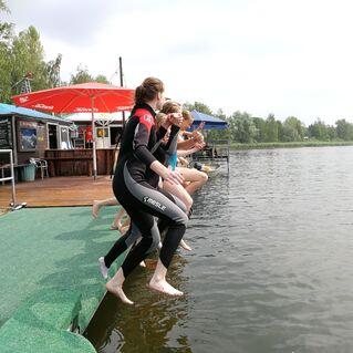 F&uuml;nf Personen in Neoprenanz&uuml;gen springen vom Steg in einen See, umgeben von B&auml;umen und einem Holzdeck.