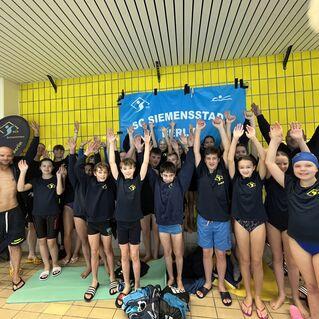 Gruppenzusammenkunft von Schwimmern in T-Shirts, alle mit erhobenen H&auml;nden, vor einer gelben Wand und einem Banner.