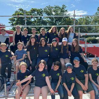 Gruppenfoto von Kindern und Erwachsenen in Sportbekleidung, die auf einem Ger&uuml;st vor einem Schwimmbad posieren.