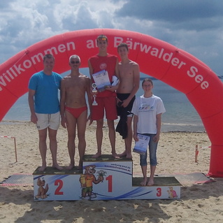 Fünf Personen stehen auf einem Siegertreppchen am Bärwalder See, umgeben von Sandstrand und Wasser.