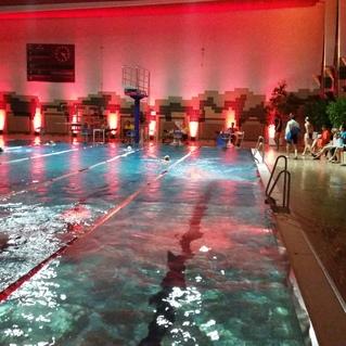 Beleuchtet Schwimmbad mit Wasseroberfläche und Schwimmern, umgeben von roten Lichtern und Zuschauern am Beckenrand.