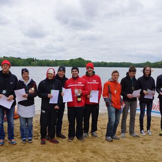 Gruppenaufnahme von zehn Teilnehmern mit Urkunden und Medaillen am Ufer eines Sees, im Hintergrund eine blaue Sportbogen.