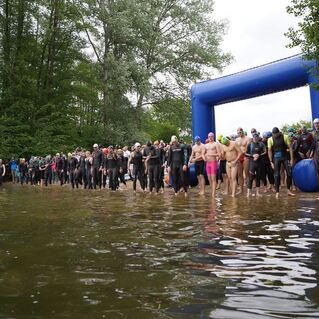 Gruppe von Wettkampfschwimmern in Neoprenanzügen am Ufer eines Gewässers, bereit für den Start.