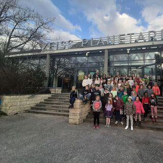 Gruppenselfie von Kindern und Erwachsenen vor dem Zeiss Planetarium, umgeben von einer grünen Landschaft und bewölktem Himmel.