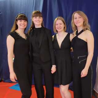 Vier Frauen in schwarzer Kleidung posieren lächelnd vor einem blauen Hintergrund auf einem roten Untergrund.