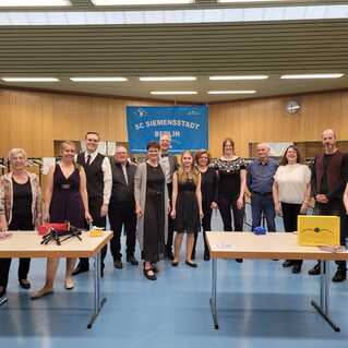 Gruppe von 12 Personen in festlicher Kleidung steht vor einer Wand mit Banner, auf Tischen sind Gegenstände und eine Spendenbox.