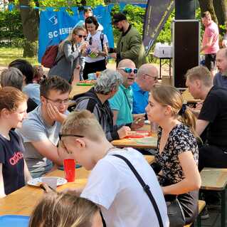Gruppe von Menschen sitzt an Tischen im Freien, isst und unterhält sich in einer grünen Umgebung.