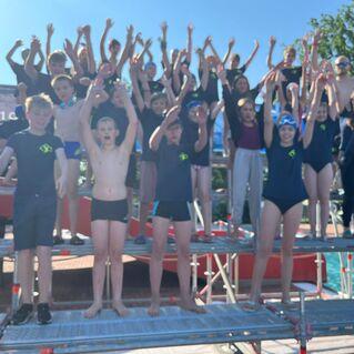 Gruppenschnappschuss von jungen Schwimmern, die auf einer Plattform stehen und ihre Arme in die Luft heben.