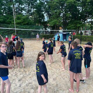 Gruppe von Kindern in blauen Shirts beim Beachvolleyballtraining an einem Sandplatz, umgeben von Bäumen.