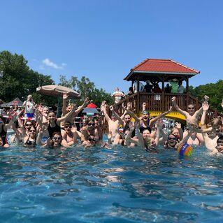 Gruppe von Kindern im Wasser, die beim Spielen mit einem Ball fröhlich die Hände heben, im Hintergrund ein Spielturm.