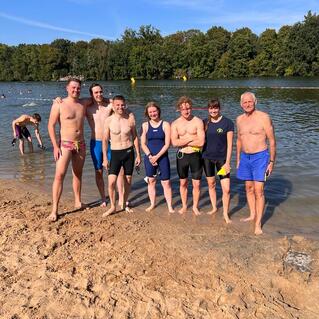 Gruppe von sieben Personen in Badebekleidung am Ufer eines Gewässers an einem sonnigen Tag.