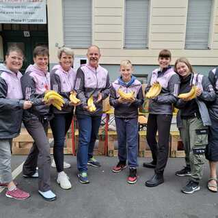 Gruppe von sieben Personen in Sportjacken hält Bananen vor Kartons auf einer Straße.
