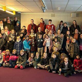 Gruppenfoto von über 50 Personen, darunter Kinder und Erwachsene, in Winterkleidung in einem hellen Raum.