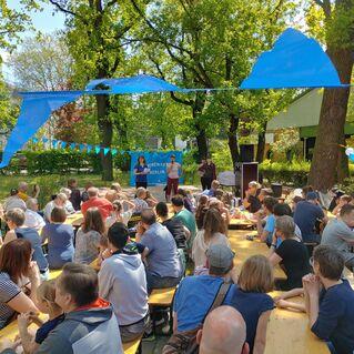 Menschen versammeln sich an Holztischen in einem Park, um einer Präsentation unter bunten Fahnen zu lauschen.