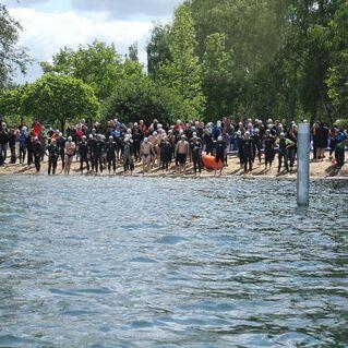 Gruppe von Schwimmern in Neoprenanzügen steht am Ufer eines Gewässers, umgeben von Zuschauern bei einer Veranstaltung.
