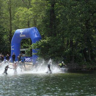 Schwimmer starten in einem Wettkampf aus einem Gewässer, umgeben von Zuschauern und Bäumen, unter einem blauen Torbogen.