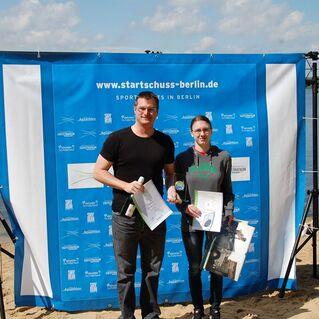 Zwei Sportler posieren mit Urkunden vor einem blauen Banner am Ufer eines Sees. Wolkiger Himmel im Hintergrund.