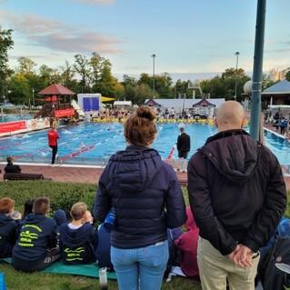 Zuschauer beobachten einen Sportwettkampf am Beckenrand eines Schwimmbads im Freien bei Sonnenuntergang.