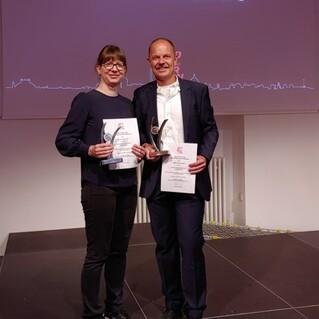 Zwei Gewinner präsentieren Pokale und Urkunden auf einer Bühne bei der Gratias-Verleihung 2023.