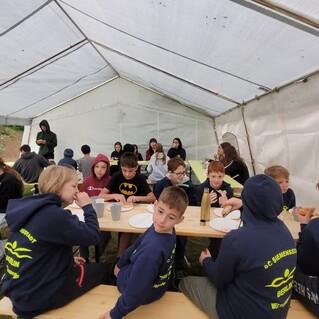 Gruppenszene im Zelt mit Kindern, die an Tischen essen und trinken, umgeben von grüner Natur.