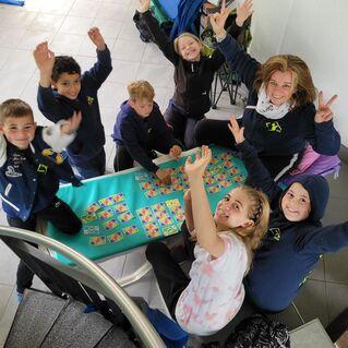 Gruppe von Kindern und Erwachsenen, die um einen Tisch mit Spielen sitzen und fröhlich die Hände heben.