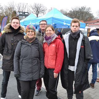 Gruppe von fünf Personen in Winterkleidung steht vor blauen Zelten auf einem Markt.