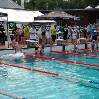Schwimmer und Trainer an einem Wettkampfpool, mit aufgehängten Werbebannern und Zuschauertribünen im Hintergrund.