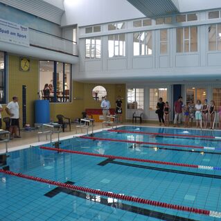 Schwimmbad mit mehreren Menschen am Beckenrand und einer Trainerin vor blauen Wasserbahnen. Zuschauer und Sportler sind zu sehen.