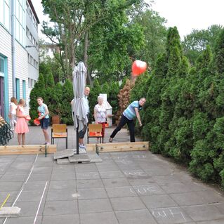 Gruppen von Personen spielen im Freien, werfen einen Ball und stehen neben einer grünen Hecke und Holzgestellen.