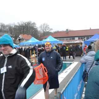 Mann in Schwimmbekleidung und Badehaube geht am Schwimmbecken vorbei, umgeben von Zuschauern und Zelten.