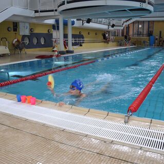 Junge schwimmt mit Schwimmhilfe in einem Hallenbad, während farbige Becher am Beckenrand stehen.