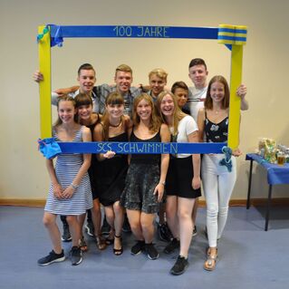 Gruppe junger Leute posiert lachend hinter einem farbigen Rahmen mit der Aufschrift „100 Jahre SCS Schwimmen“.