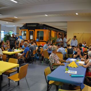 Blick in einen Festsaal mit Gästen an bunten Tischen und einem orangefarbenen Zug im Hintergrund.