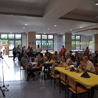 Menschen stehen und sitzen in einem festlich dekorierten Raum mit blauen und gelben Girlanden und Tischen.