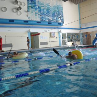 Zwei Personen im Wasser mit Schwimmreifen und Paddel, Zuschauer stehen am Beckenrand in einem Hallenbad.