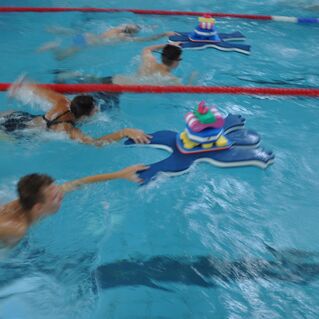 Gruppenschwimmen von Kindern, die auf bunten Schwimmmatten im Wasser paddeln, während Wellen spritzen.